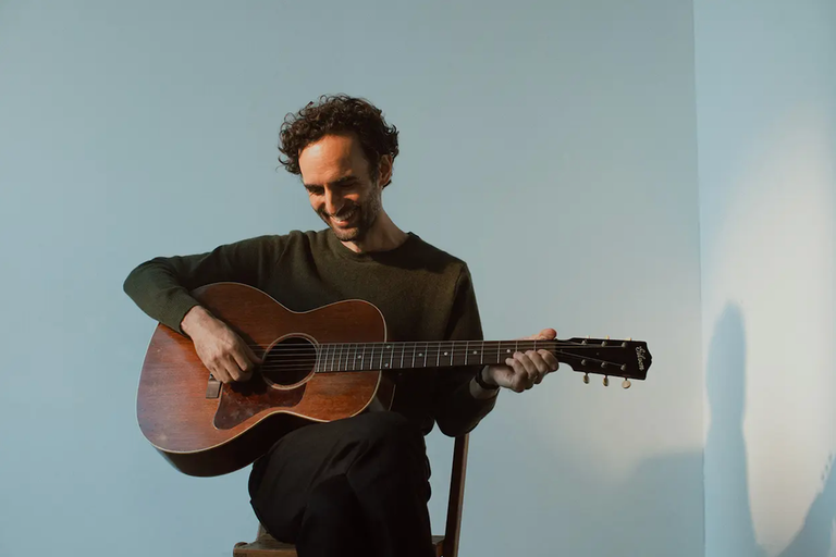 Julian Lage playing acoustic guitar while sitting on a chair smiling against a plain backdrop, photographed by Alysse Gafkjen.